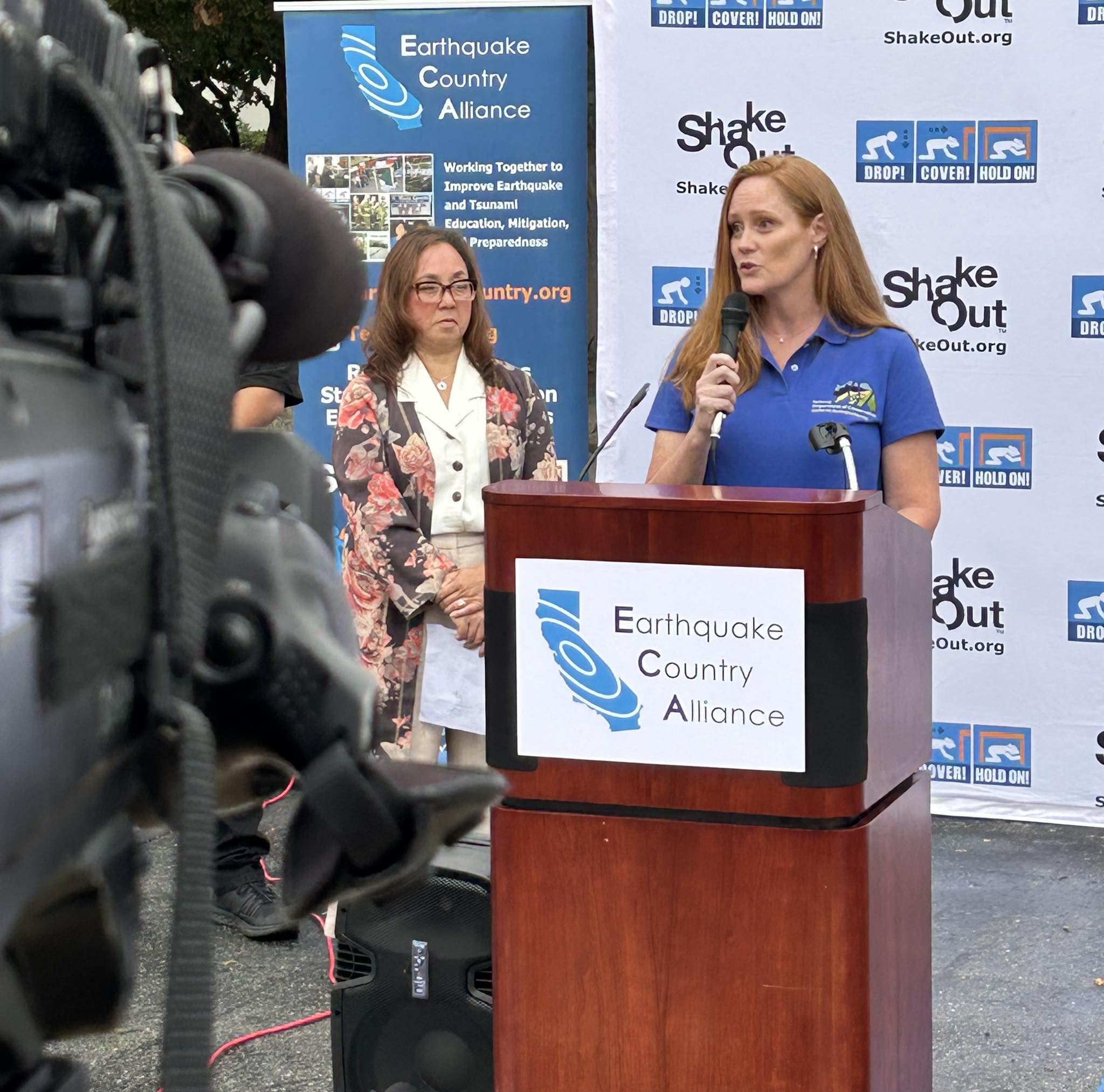 Wendy in a blue shirt, holding a microphone at a ShakeOut podium.