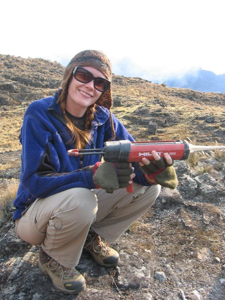 Wendy Bohon smiling in the field, wearing a blue fleece and holding a red rock-sampling drill.