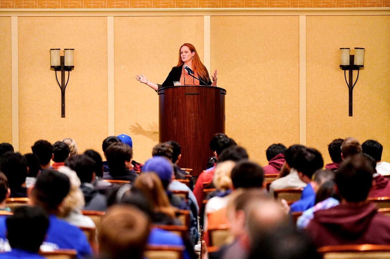 Wendy speaking at a podium, with a large audience in the foreground.